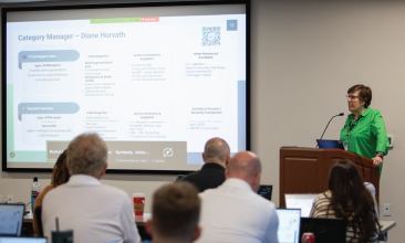 VITA procurement professional standing at a podium facing a room full of event attendees. Screen features a slide explaining the IT procurement process for the state.